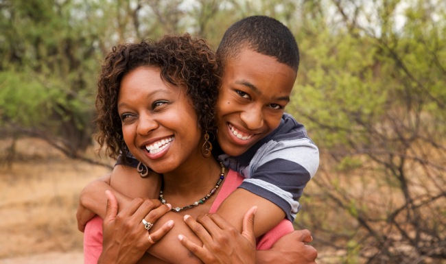 son hugs his mom from behind as they smile in the park